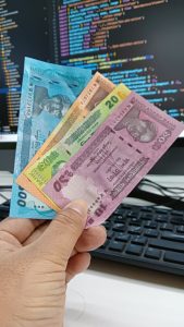 A hand is holding three banknotes of different denominations: a blue 100 taka bill, an orange 20 taka bill, and a pink 50 taka bill. The background features a computer screen displaying lines of code