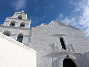 Low angle view of the front of Mission San Diego de Alcala. An old church with white stucco with a bell tower and cross against a bright blue sky.