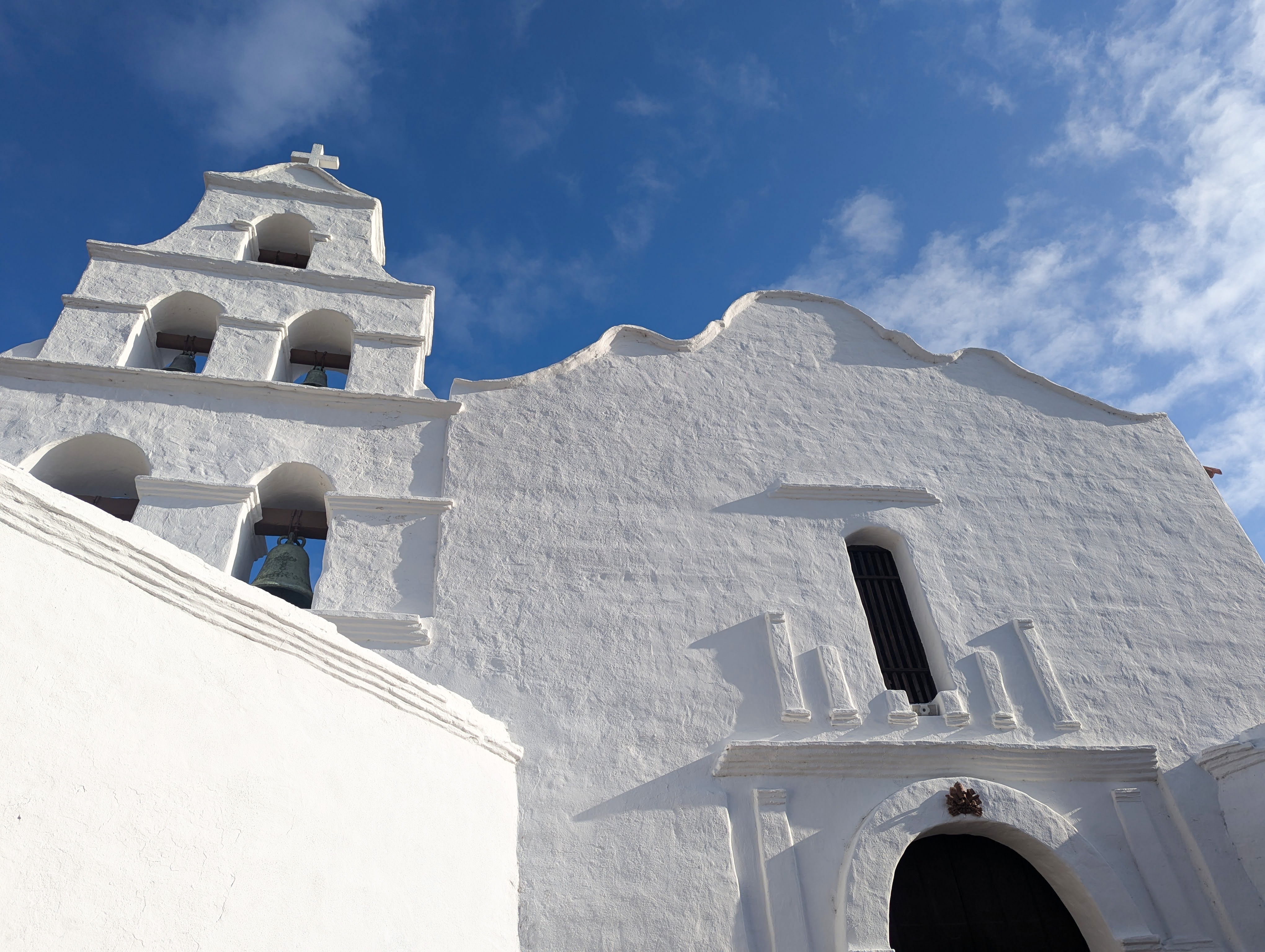 Low angle view of the front of Mission San Diego de Alcala. An old church with white stucco with a bell tower and cross against a bright blue sky. 