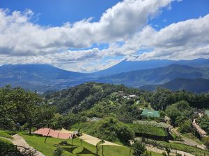 Vista panorámica de un paisaje montañoso con cielo azul y nubes blancas, colinas cubiertas de vegetación y un valle en el horizonte, con áreas verdes y estructuras de descanso en primer plano. 
