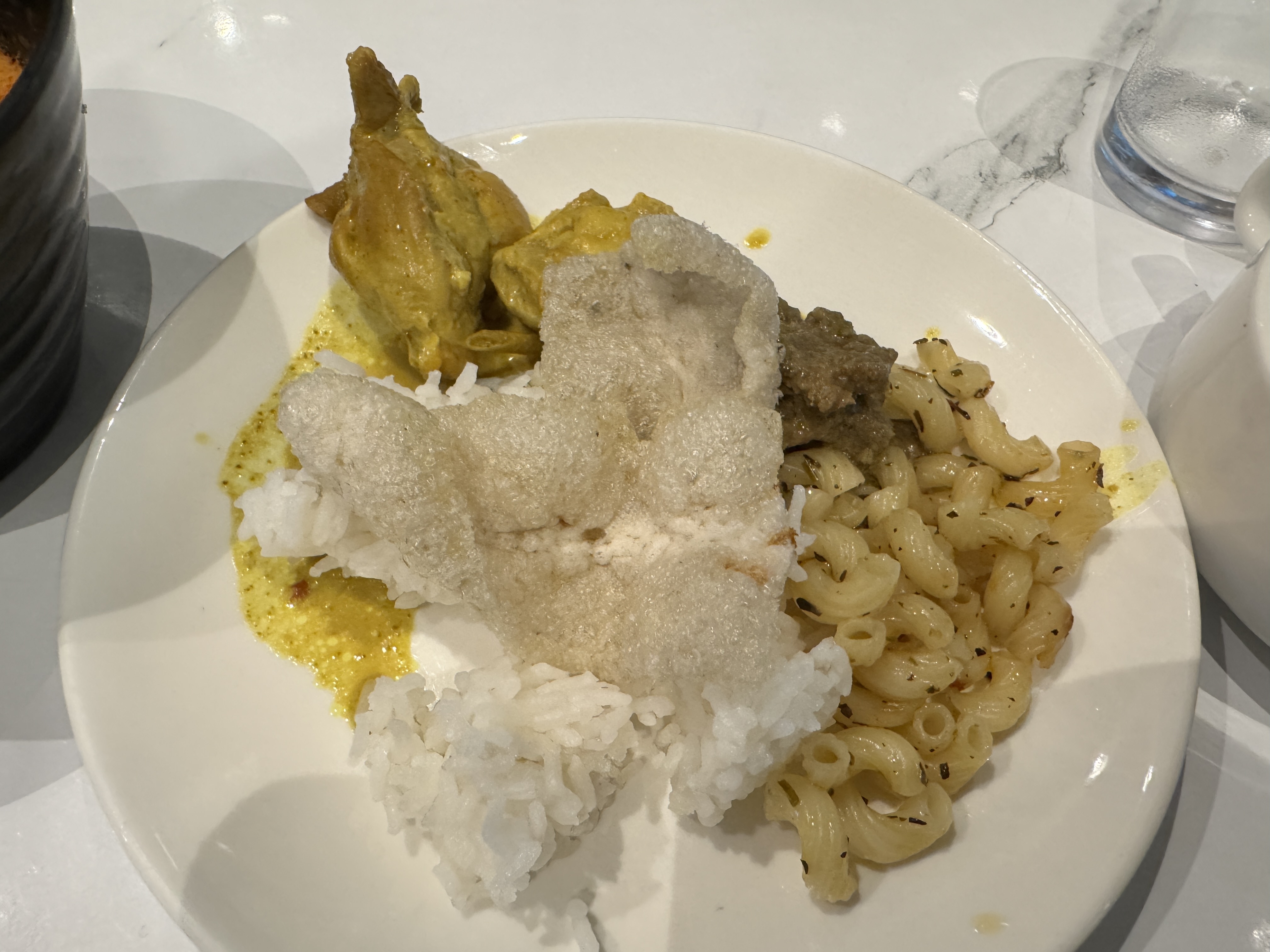 On a white plate: elbow macaroni, white rice, yellow chicken curry, and beef, topped with a big, airy rice cracker.