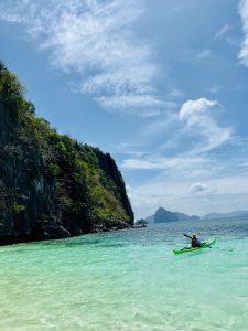 Kayaker paddles in crystal-clear turquoise waters near a lush, rocky cliff under a vibrant blue sky.