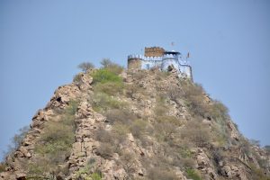 A hilltop temple fortress with white and blue walls rises dramatically above rocky slopes and sparse vegetation, viewed from Moosi Maharani Ki Chhatri, Alwar, Rajasthan.
