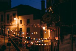 Nighttime street scene in Porto during the summertime festival São João, with colorful string lights and castanets - traditional percusion instruments - hanging across the narrow street. Old buildings are softly lit by warm street lamps, and other traditional festival decorations hang in the foreground, slightly out of focus.