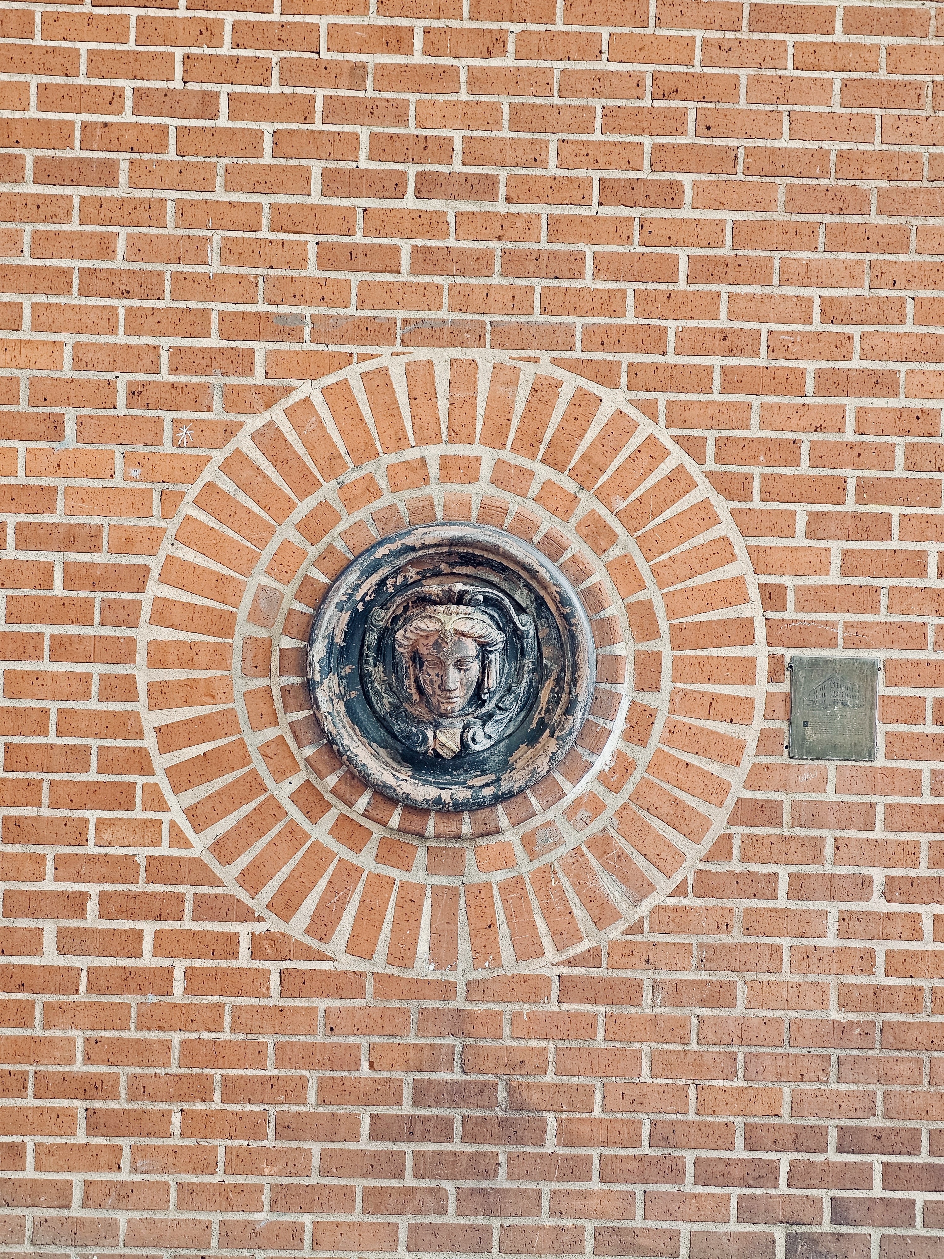 A decorative medallion featuring a sculpted face, encircled by concentric patterns of red bricks, set into a wall in Portland, Oregon.