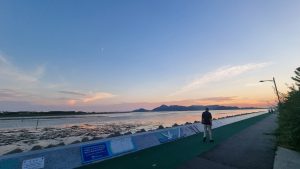Sunset view at the Nakdong River Estuary Wetland Protected Area near Myeongji Bird Observatory, with people walking along the riverside path and distant mountains silhouetted against the colorful evening sky
