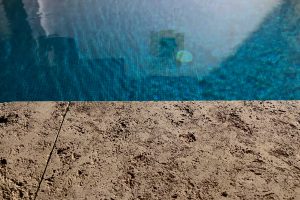 Edge of a sand-colored stone pool in the lower half, with the blue water and a patterned wall in the background