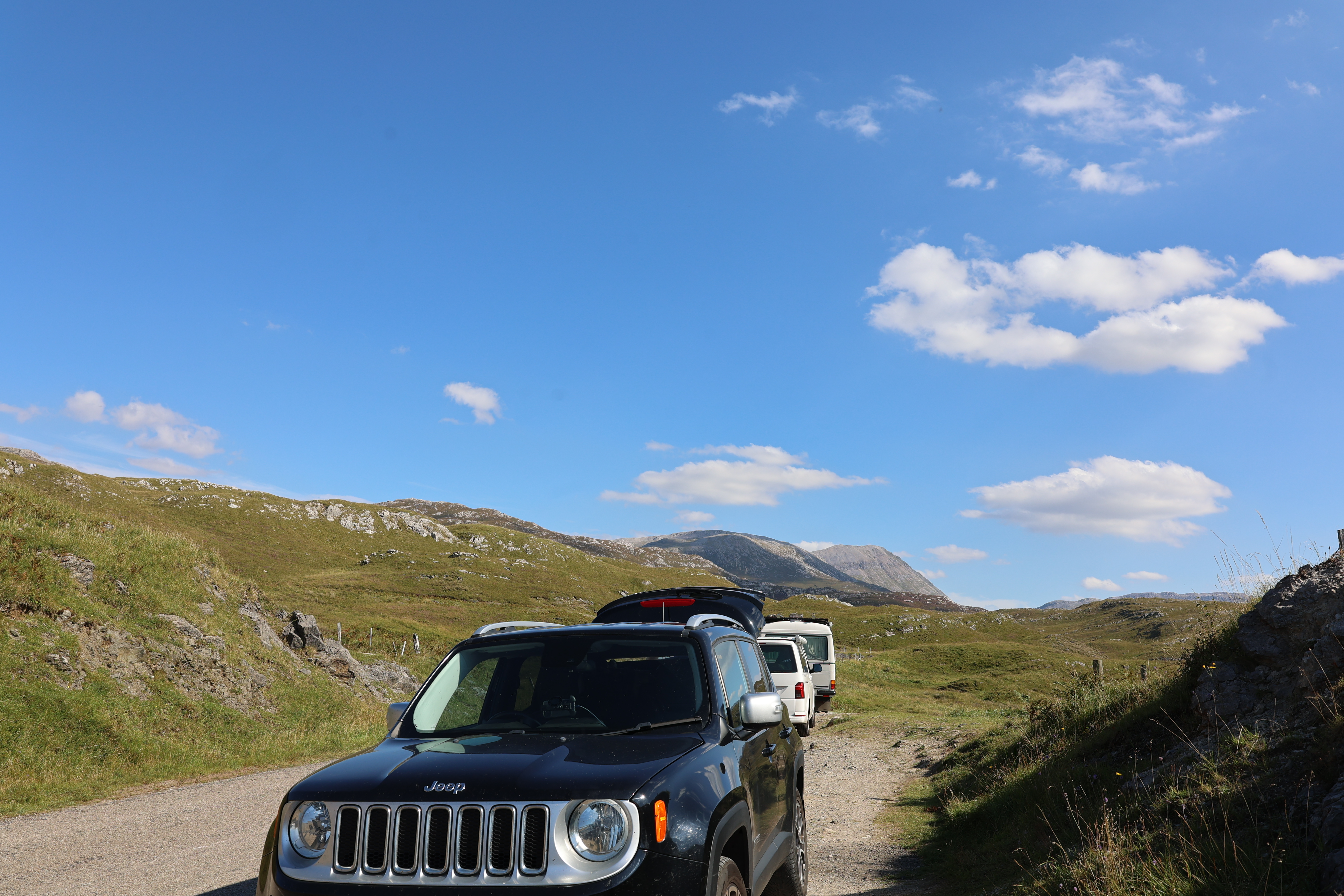 A black Jeep sits on a gravel road amid green hills and rocky terrain under a sunny, cloud-dotted Scottish sky