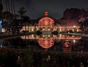 A night time view of the botanical building in Balboa Park in San Diego. The building is surrounded by palm trees and is reflected in the pond.