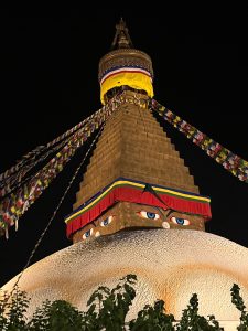A close-up view of a Buddhist stupa illuminated at night, showcasing its intricate architecture. The structure features a prominent golden spire, vibrant prayer flags cascading down, and large, expressive eyes depicted on the sides of the stupa. The base is rounded and textured, surrounded by some greenery. The scene conveys a sense of serenity and cultural significance.