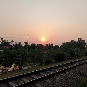 The image captures a serene sunset view taken from a moving train, with railway tracks in the foreground and a soft orange sun descending above a line of silhouetted trees and houses. The sky is hazy, creating a warm glow, while faint reflections of lights on the window glass suggest the photo was taken indoors, likely through a train window. The mood is peaceful and contemplative, evoking the quiet charm of rural travel at dusk. #SummerPhotoContest