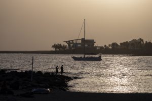A serene sunset view from Playa de Las Vistas in Playa de las Américas, Tenerife, with a sailboat on calm waters, two silhouetted figures standing on the rocky shore, and modern buildings and palm trees in the background.