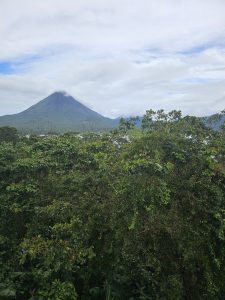 Panoramic view of Arenal Volcano partially covered by clouds, surrounded by lush tropical vegetation under a cloudy sky.
