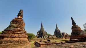 A low-angle photo showing the ruins of several large, cone-shaped stone temples in Ayutthaya, Thailand, with a clear blue sky in the background.