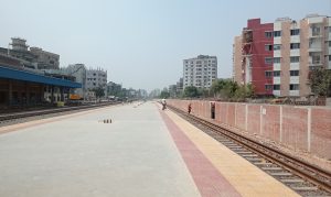 A view of a railway station under a clear blue sky, featuring empty platforms lined with yellow and red tiles. On one side, there is a blue-roofed station building and several tall buildings in the background.