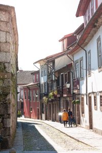 Two people walking along a narrow cobblestone street lined with colorful historic houses and balconies in Porto, Portugal, on a sunny day. 