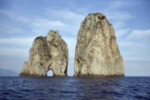 I Faraglioni, Capri — towering limestone sea stacks with warm-toned cliffs, one pierced by arches, rising from the Tyrrhenian Sea under a pale blue sky.