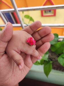 A hand gently holds a small pink flower resting on a child's hand. The background features a vibrant yellow wall and green plants