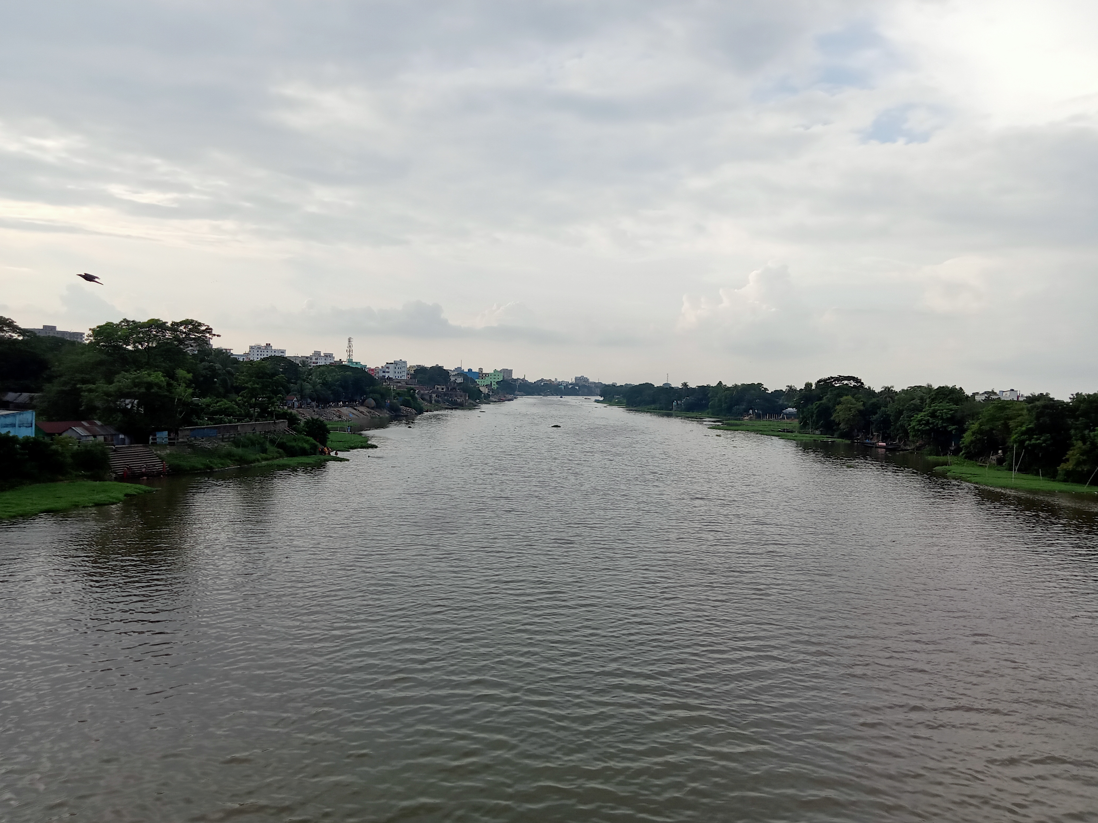 Titas River flows through a landscape lined with greenery and urban structures under a cloudy sky