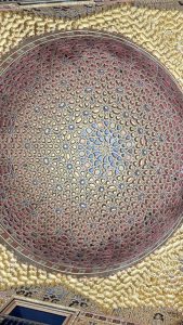 A beautifully ornate ceiling featuring intricate geometric patterns in gold, red, and blue. The design includes star-like shapes and is highlighted by a dome structure that adds depth and elegance to the space