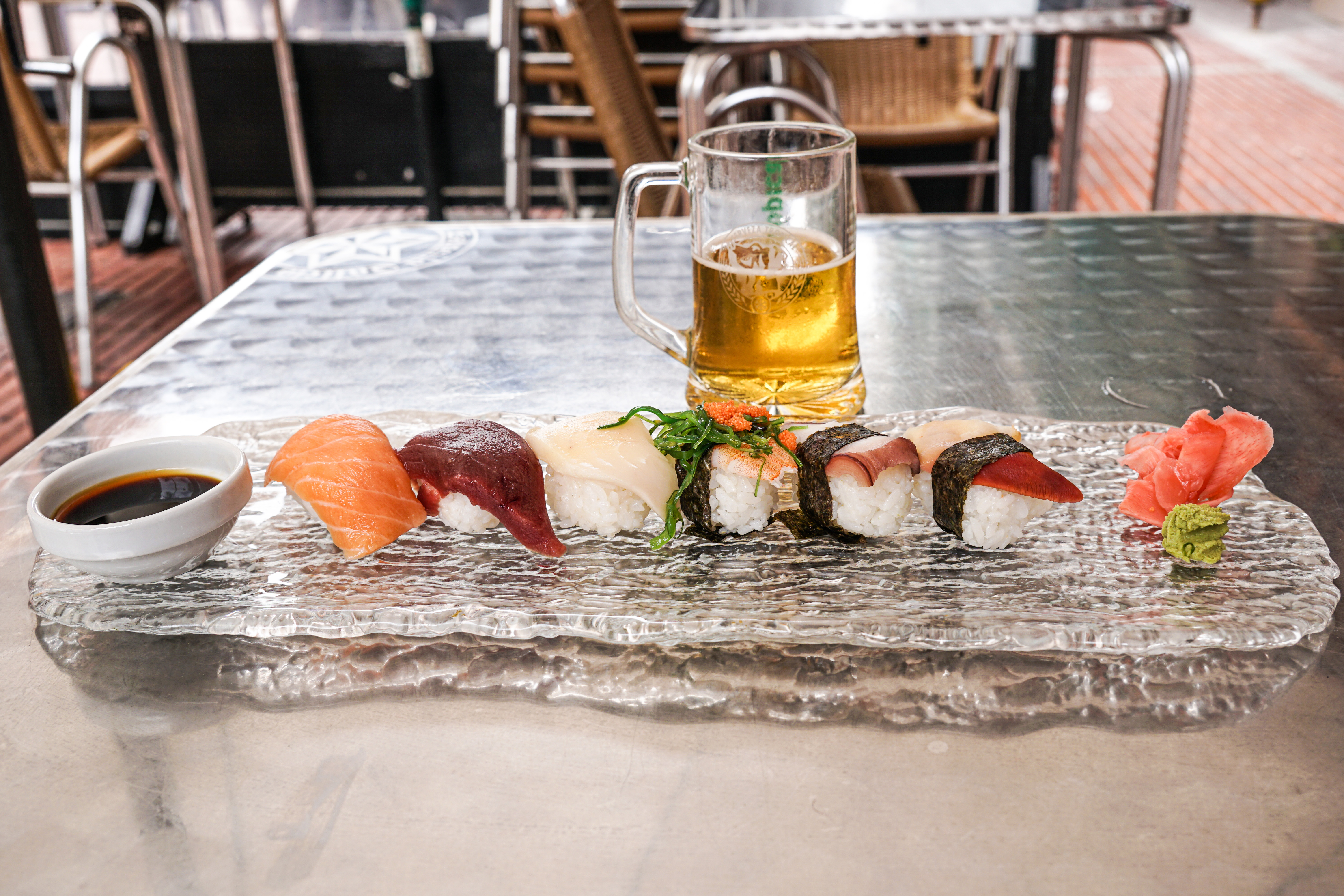 A plate of sushi is beautifully arranged on a textured glass serving dish. The sushi includes various types such as salmon, tuna, and white fish, with some pieces wrapped in nori. Accompanying the sushi is a small bowl of soy sauce, a dollop of wasabi, and pickled ginger.