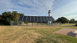 A solar panel array on a lawn.  A lighthouse pokes up above in the distance behind.