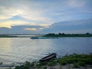 A serene river scene at dusk, featuring a small wooden boat resting on the shore amidst patches of green vegetation. In the background, a larger boat navigates the water, surrounded by gentle waves reflecting the colors of the sky. Power lines and distant trees line the waterway, creating a tranquil yet industrial atmosphere.