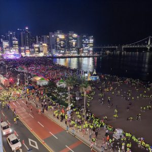 A large crowd gathered at Gwangalli Beach in Busan for a night festival, with the illuminated Gwangan Bridge in the background. 2025 NIGHT RACE IN BUSAN