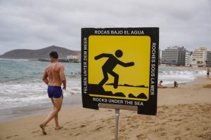 A beach scene featuring a warning sign that reads "Rocks Under the Sea" in multiple languages, including Spanish, German, and French. The sign displays an image of a person slipping on rocks. In the background, a man walks along the shoreline wearing swim shorts, while other beachgoers enjoy the water. 