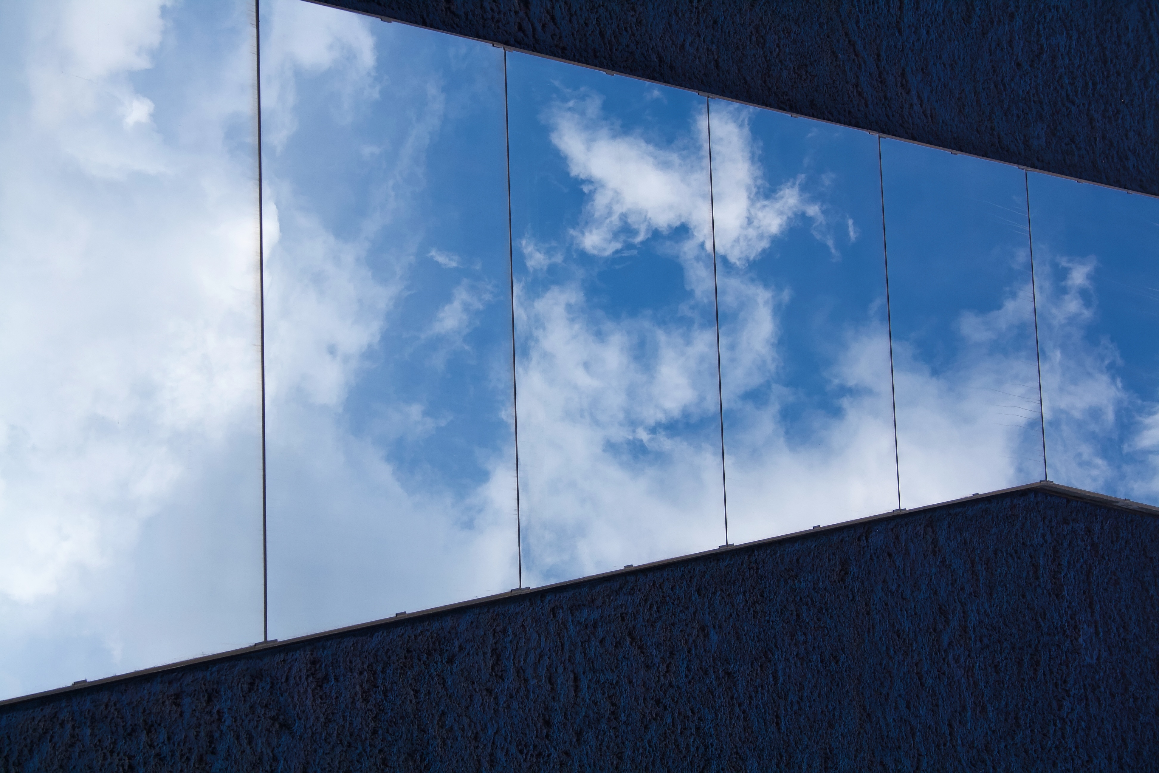 Cloudy blue sky reflected in geometric mirrors on a dark blue stucco wall.