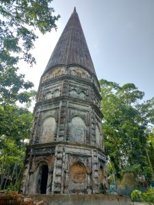 A weathered, conical tower with a pointed top rises amidst lush greenery. 
