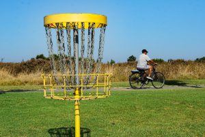 A yellow disc golf basket with chains stands in the foreground on a grassy field. In the background, a man rides a bicycle along a pathway, surrounded by tall grass and shrubs under a clear blue sky. 