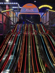 A vibrant indoor play area featuring three shiny black slides with colorful neon stripes in red, orange, blue, and yellow. Above the slides, there is a large sign with a cartoonish car and the text “HELLO THE HOPUP” and “HAPPY HOPUP TIME,” set against a dark ceiling adorned with bright lights
