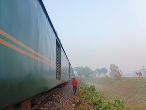 A long green train car with yellow stripes is parked beside a railway track, surrounded by a misty landscape. A person in a red jacket walks alongside the train