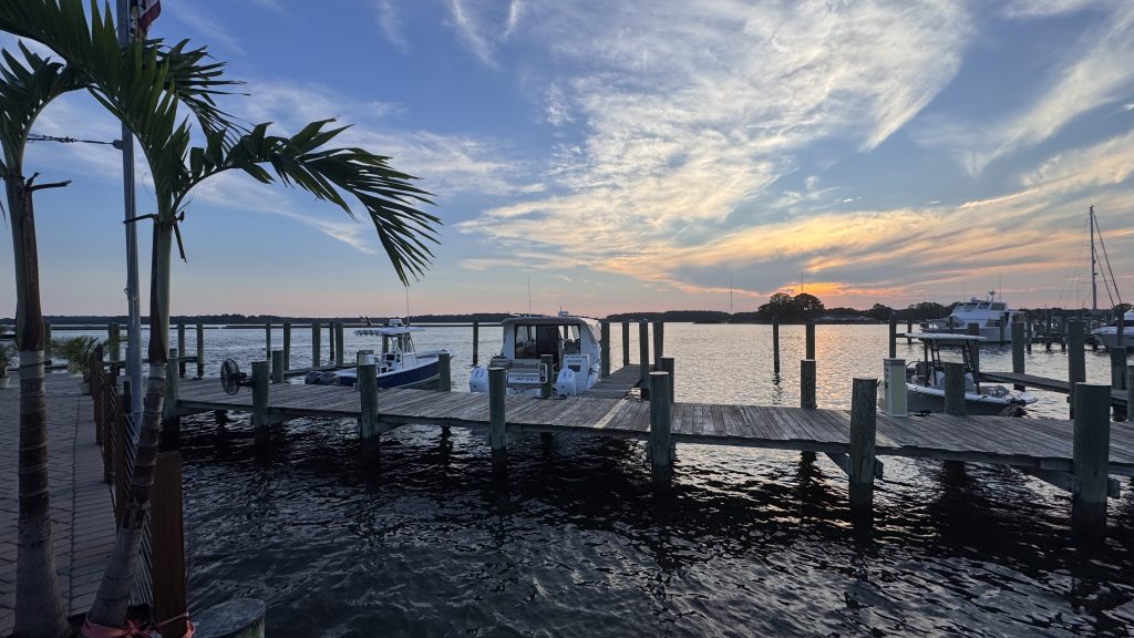 Docks at Kent Island Narrows Maryland, featuring several boats moored alongside a wooden dock. Palm trees are visible in the foreground, with a clear sky showcasing vibrant colors as the sun sets on the horizon