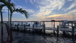 Docks at Kent Island Narrows Maryland, featuring several boats moored alongside a wooden dock. Palm trees are visible in the foreground, with a clear sky showcasing vibrant colors as the sun sets on the horizon