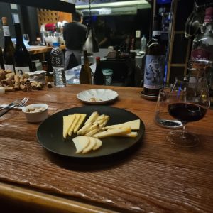 A wooden table is set with a variety of cheeses arranged on a black plate, accompanied by a small dish of nuts. A glass of red wine sits nearby, along with a bottle of wine and a couple of empty glasses. In the background, blurred figures are seen working in a kitchen, with bottles of wine and corks scattered around the bar area
