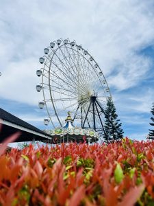Ferris wheel against a blue sky, framed by red flowers and a carousel below. The scene conveys a cheerful, playful atmosphere with a sense of leisure.