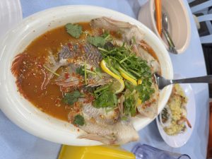 A white oval platter filled with a whole steamed fish, garnished with fresh cilantro, thin slices of lemon, and chopped green onion. The fish is surrounded by a rich, spicy sauce containing red chili flakes and herbs. In the background, a small plate with a serving of fried rice and seafood can be seen, along with a bowl of soup and a pair of chopsticks. The setting features a blue table surface.