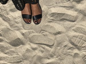 A pair of feet wearing black sandals stands on a sandy beach.