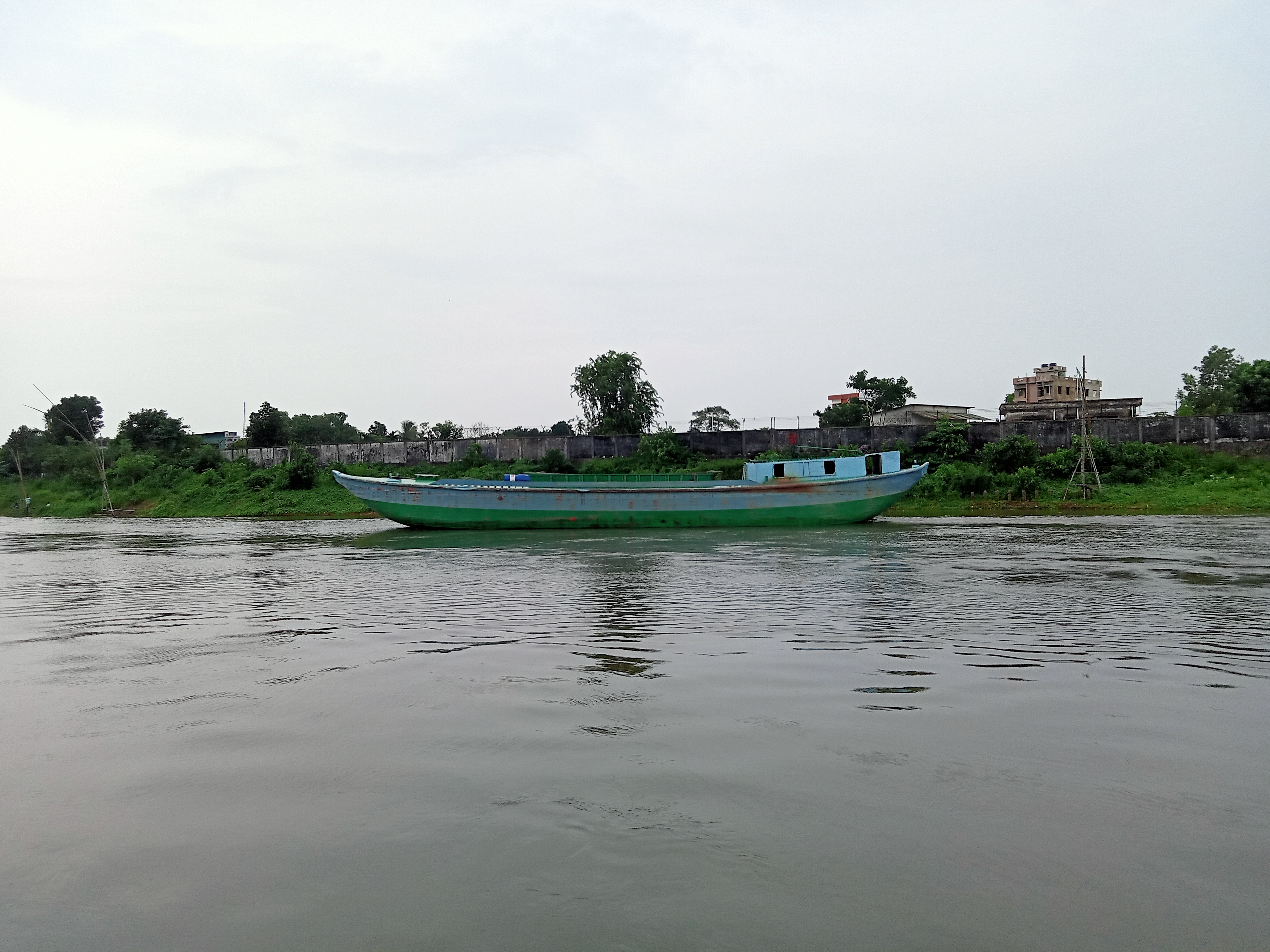 A wooden boat painted in shades of green is anchored in a calm river, with trees and lush greenery lining the opposite bank. In the background, a low stone wall and a few buildings are visible