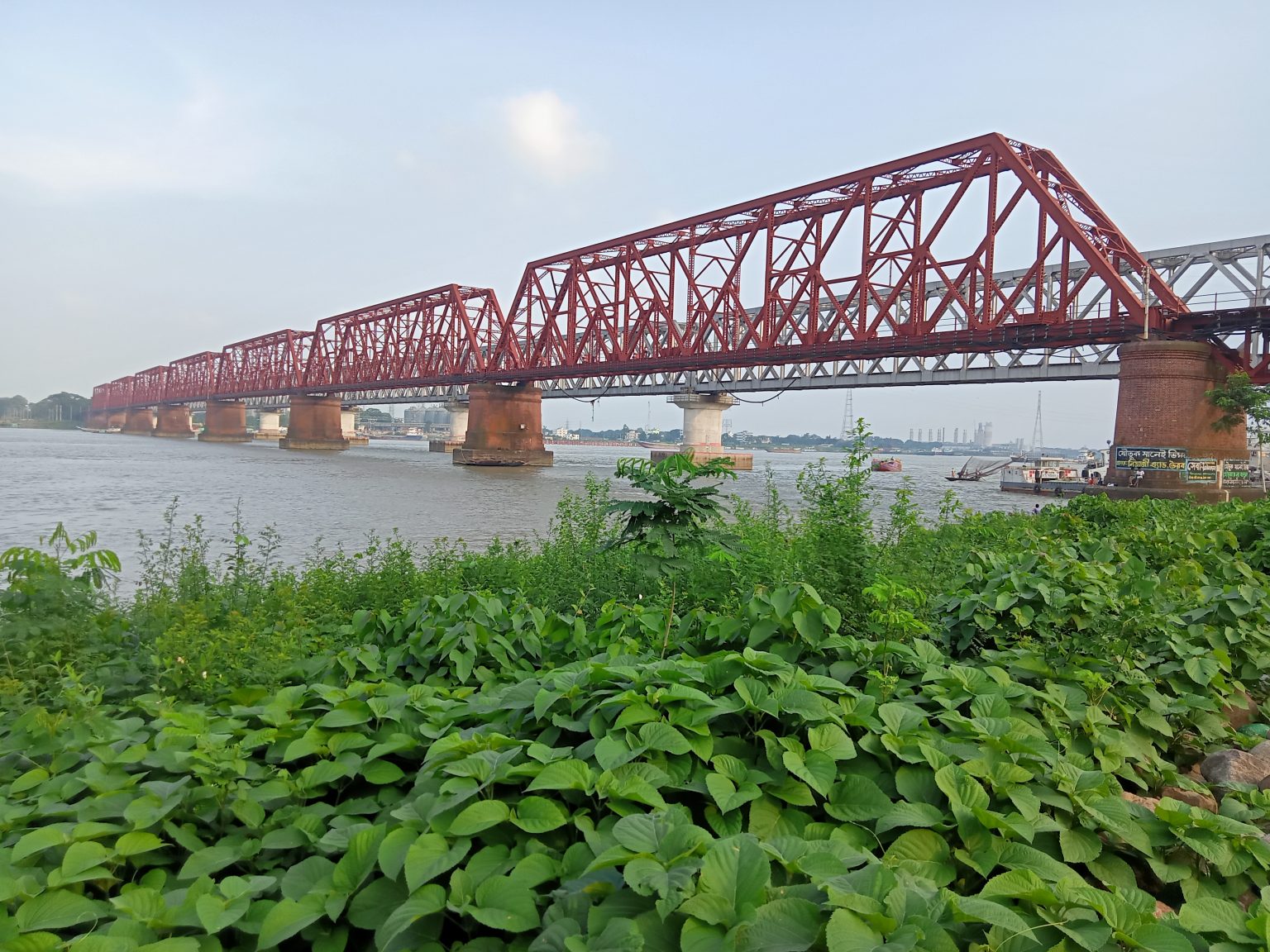A picture of a red coloured bridge which is supported by big cemented stands in between, underneath is a river. Bhairab, Kishoreganj, Bangladesh