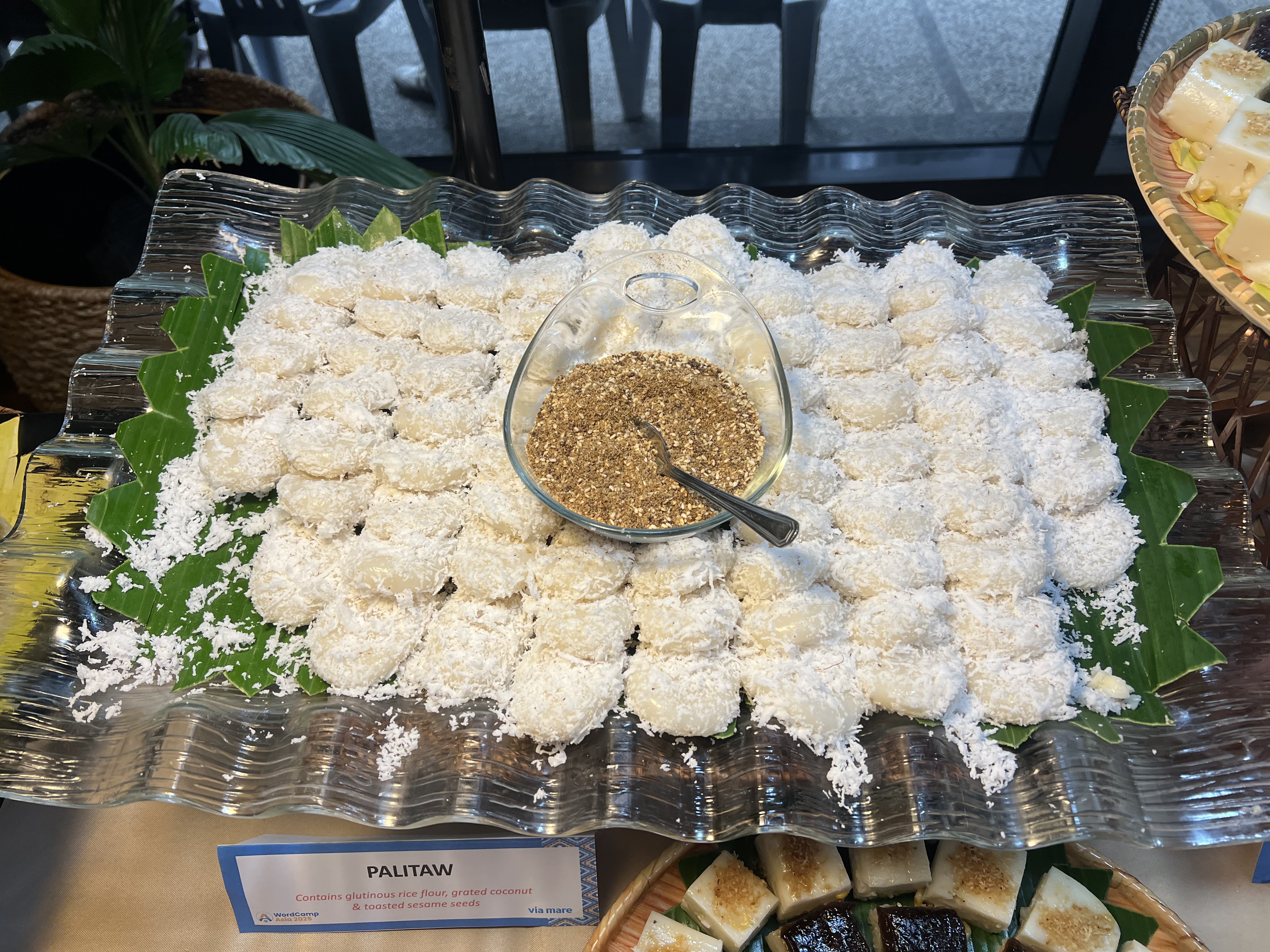 A large platter of small, white oval-shaped desserts called Palitaw, covered in grated coconut, with a separate bowl of brown sugar and sesame seeds in the center.