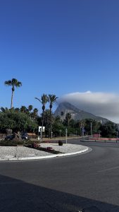 A scenic view of Gibraltar with a roundabout of white pebbles, colorful flowers, palm trees, and the cloud-topped Rock of Gibraltar in the background.