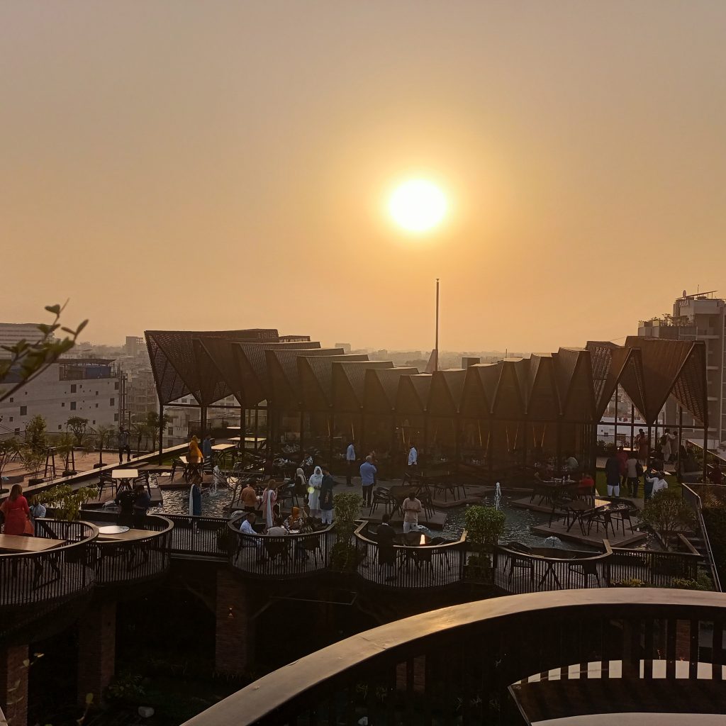 A sunset view from a rooftop terrace, showcasing a warm golden sun in the clear sky. The terrace features modern architectural elements with wooden structures and seating areas. People are gathered, engaging in conversations, while plants and greenery provide a lively atmosphere. In the background, the city skyline is visible, enhancing the urban setting.