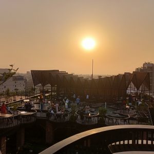 A sunset view from a rooftop terrace, showcasing a warm golden sun in the clear sky. The terrace features modern architectural elements with wooden structures and seating areas. People are gathered, engaging in conversations, while plants and greenery provide a lively atmosphere. In the background, the city skyline is visible, enhancing the urban setting.

