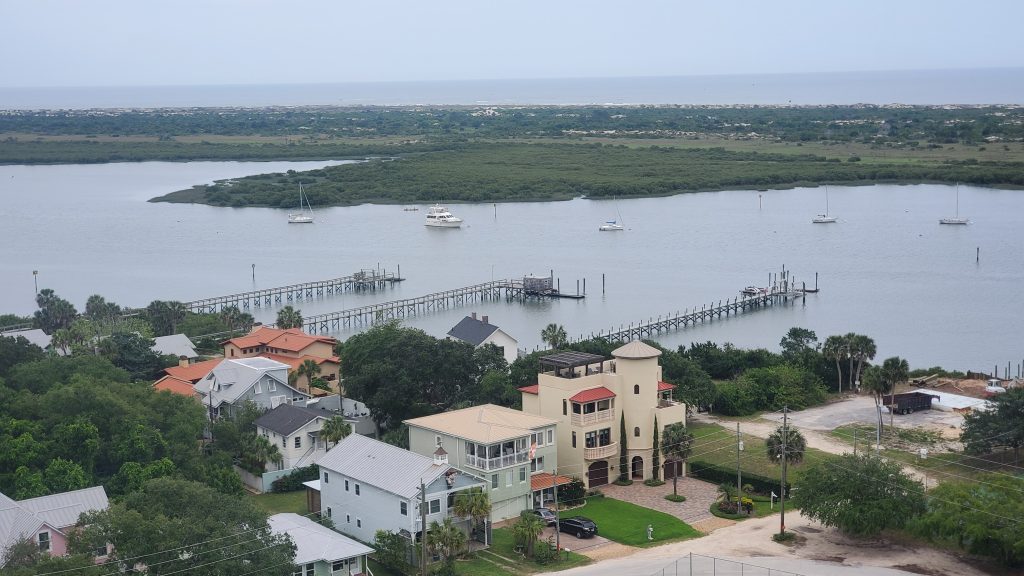 View looking down from a lighthouse that overlooks greenary, trees and bushes, house buildings and boat docks, a cove and a few boats, cars on Florida’s East Coast.