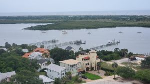 View looking down from a lighthouse that overlooks greenary, trees and bushes, house buildings and boat docks, a cove and a few boats, cars on Florida’s East Coast.