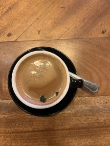 An overhead shot of a black and white coffee mug on a black saucer with a spoon. The mug is full of a hot coffee drink with a small layer of foam. The mug is sitting on a wooden table