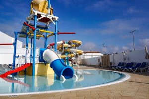 A colorful water play structure stands next to a clear, shallow pool. The structure features a large blue slide and several water slides of varying heights in shades of yellow, red, and beige. Surrounding the pool are several blue sun loungers and umbrellas 